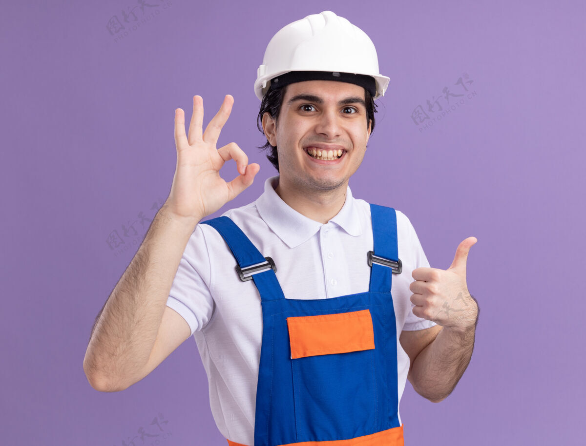 年轻身穿施工制服 头戴安全帽的年轻建筑工人站在紫色的墙上 高兴而积极地看着前方 露出ok的手势 竖起大拇指好建设者快乐