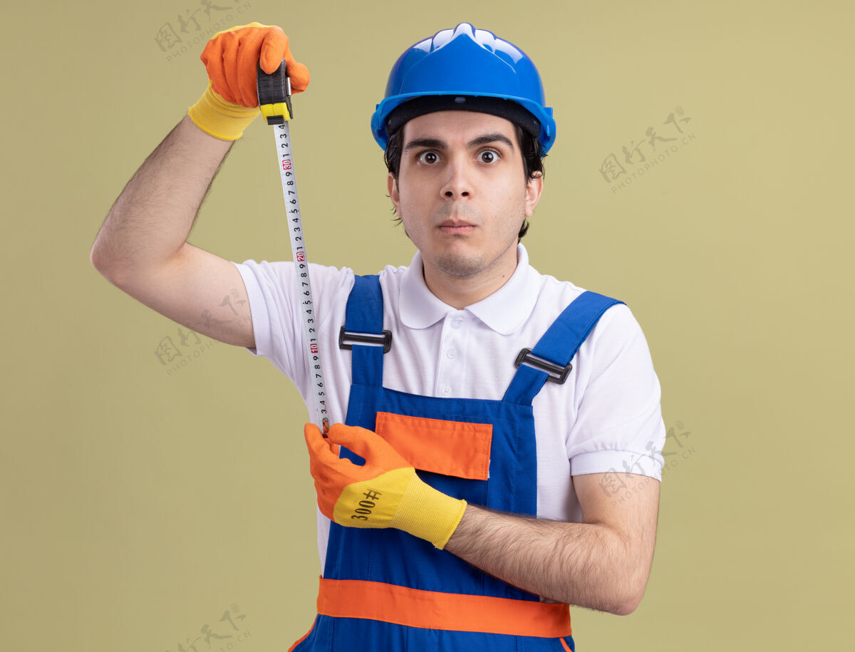 建筑工人年轻的建筑工人穿着建筑制服 戴着安全帽 戴着橡胶手套 手里拿着卷尺 站在绿色的墙壁上困惑地看着前方制服持有胶带