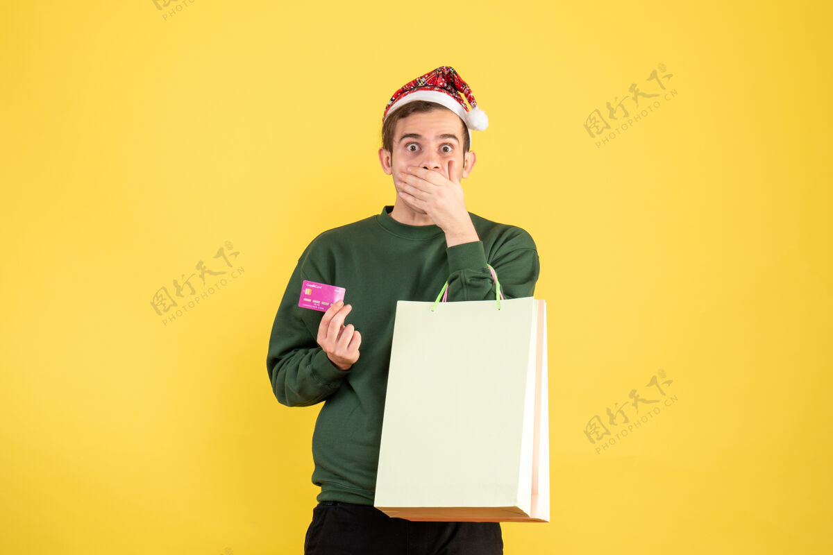 人正面图：戴着圣诞帽的年轻人拿着购物袋和卡片 手放在黄色背景上的嘴巴上秘书圣诞节举行