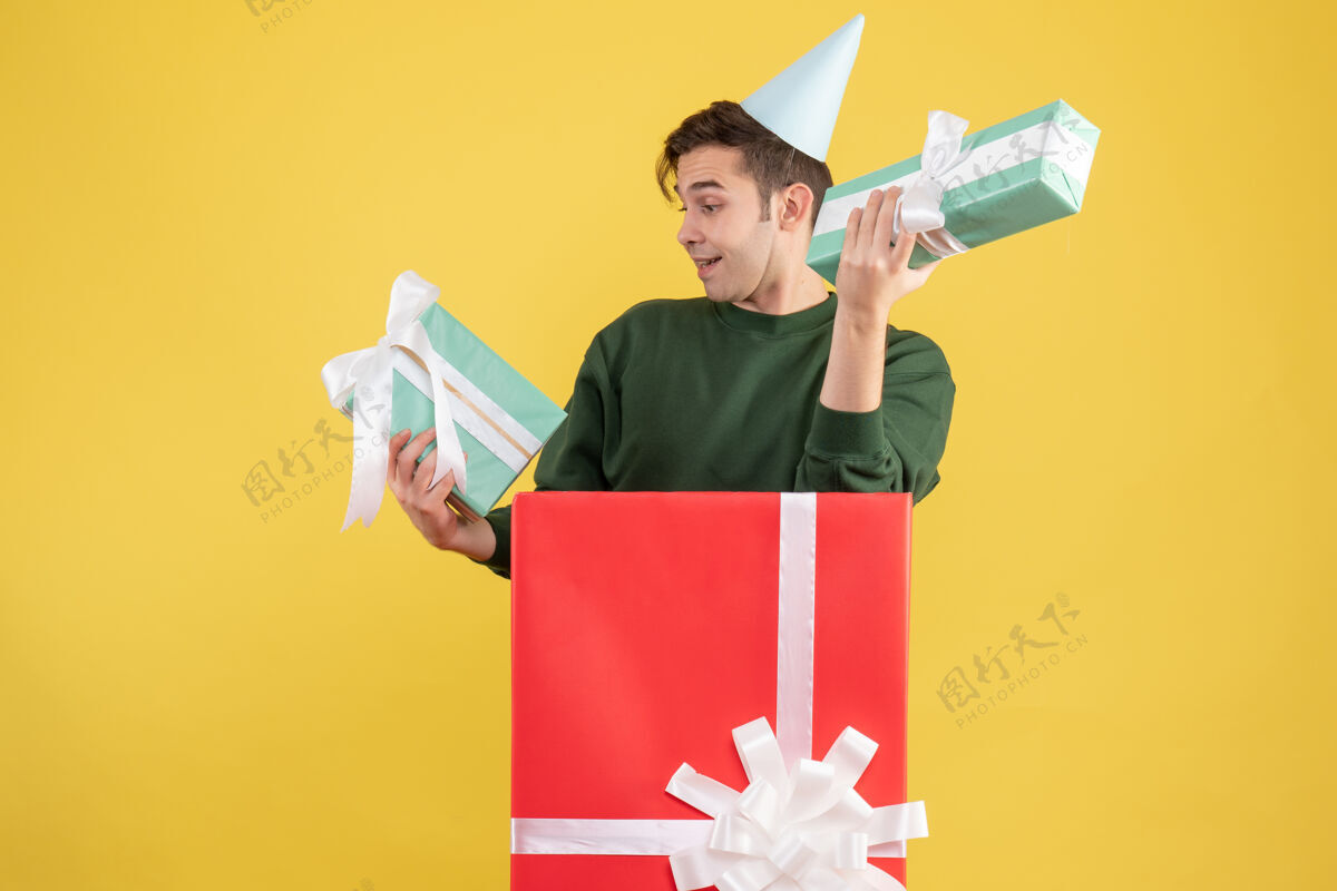 快乐正面图：戴着派对帽和礼物的年轻人站在黄色背景的大礼盒后面肖像大站立