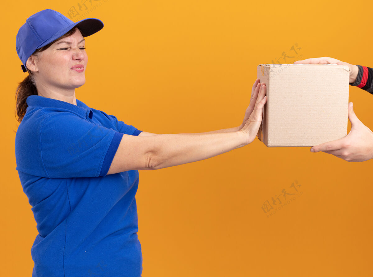 送货身穿蓝色制服 戴着帽子的年轻送货员站在橘色的墙上 表情厌恶 满脸忧虑 拒绝拿包裹包装帽子站着