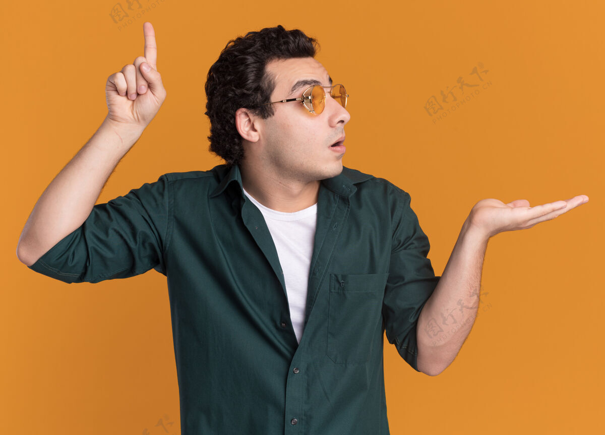 惊喜身穿绿色衬衫 戴眼镜的年轻人一边看着一边惊讶地用食指指着 一边举着手臂 一边站在橙色的墙上介绍手臂站着