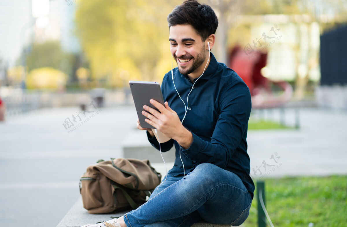 男性年轻人坐在户外的长椅上通过数码平板电脑进行视频通话的照片城市概念触摸屏城市休闲