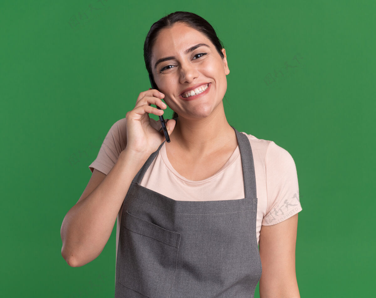 手机站在绿色的墙上 穿着围裙的年轻美发师微笑着看着前面讲着手机围裙微笑年轻
