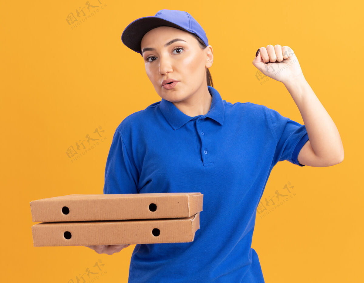 制服身穿蓝色制服 头戴鸭舌帽的年轻女送货员拿着披萨盒 自信地看着前方 举起拳头站在橙色的墙上拿着盒子女人