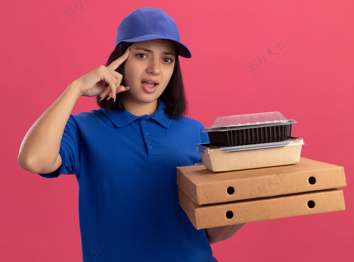 指着身穿蓝色制服 头戴鸭舌帽的年轻送货女孩拿着披萨盒和食品包 用食指指着太阳穴 神情迷茫 忘了站在粉红色的墙上帽子拿着忘了