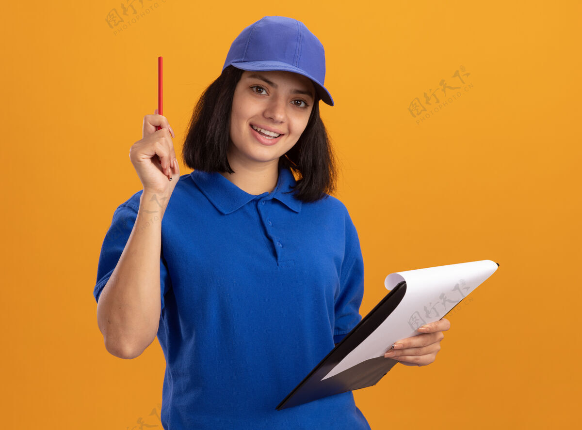微笑身穿蓝色制服 戴着帽子 拿着空白页和铅笔的剪贴板的年轻送货女孩站在橙色的墙上 开心而积极地微笑着制服铅笔快递
