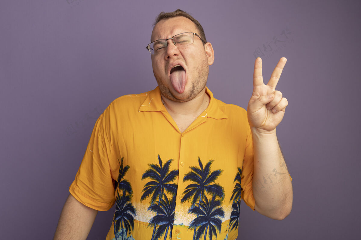 坚持一个戴着眼镜的男人穿着橘色衬衫 伸出舌头 在紫色的墙上站着一个快乐的v型标志欢乐穿着舌头