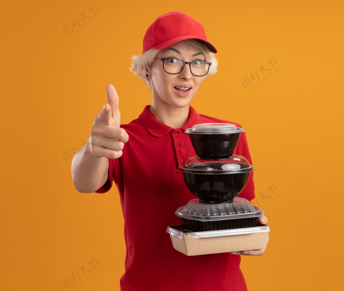 穿着身着红色制服 戴着帽子 戴着眼镜 拿着一叠食品包 用食指指着 微笑着自信地站在橙色的墙上 快乐的年轻女送货员目录年轻人帽子