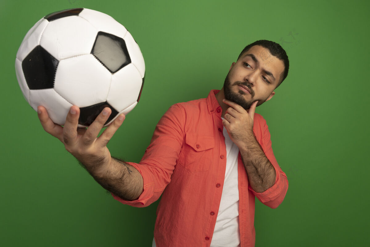 思考身穿橘色衬衫 留着胡子的年轻人拿着足球 站在绿色的墙壁上 看着它 好奇地思考着球阴谋男人
