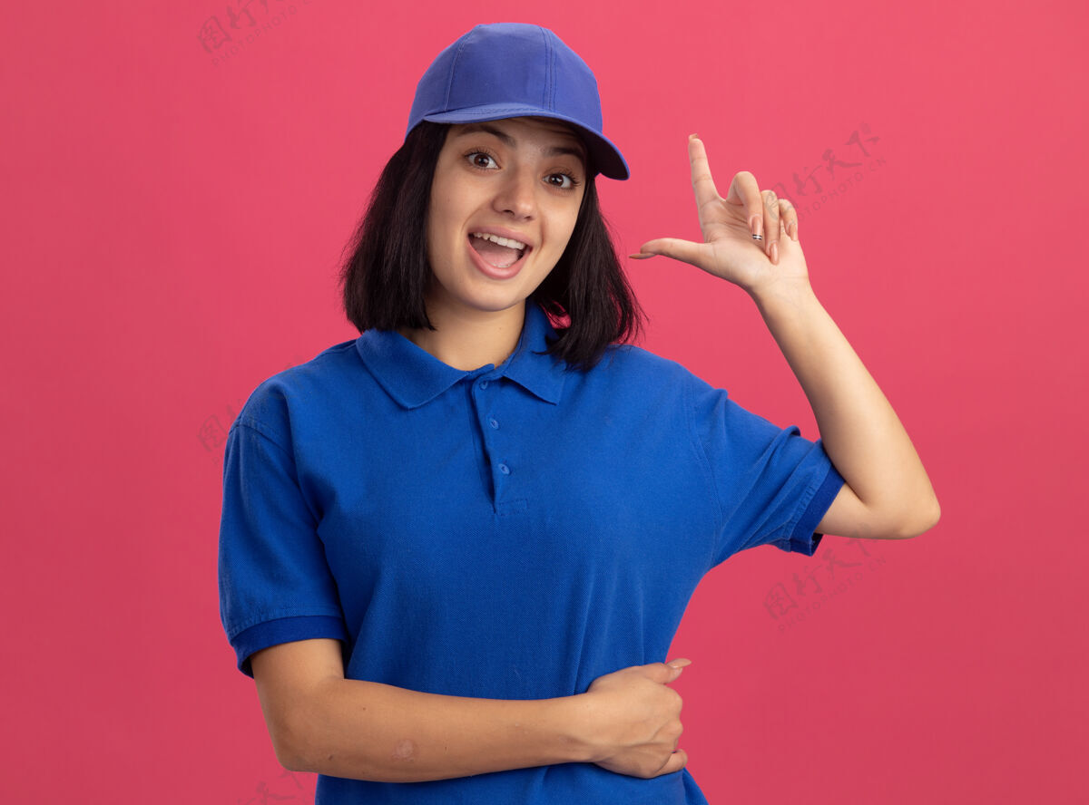 手指身穿蓝色制服 头戴鸭舌帽的年轻送货小姐站在粉红色的墙上 高兴而又惊讶地展示着有新想法的食指女孩惊喜制服
