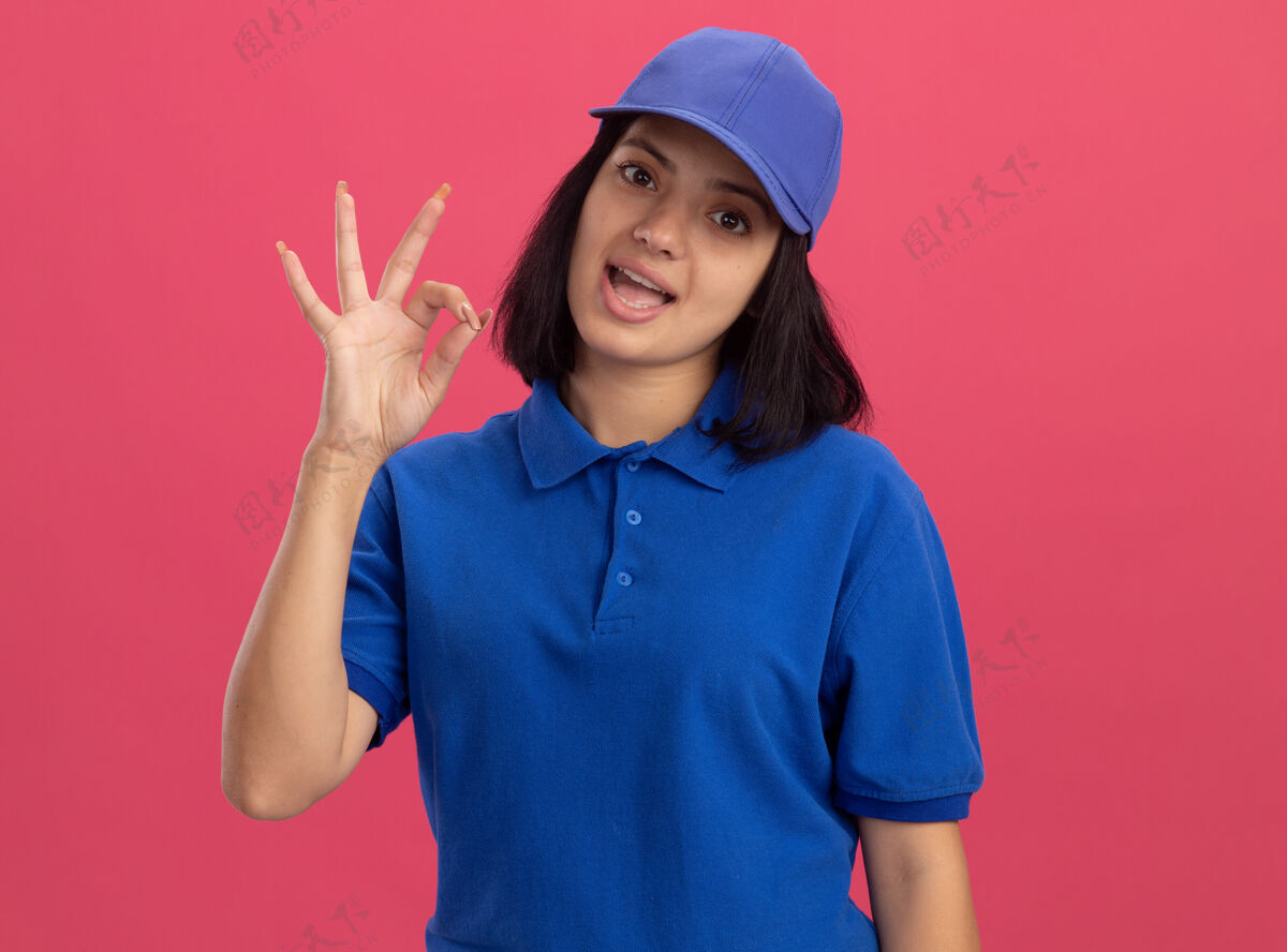 好身穿蓝色制服 头戴鸭舌帽的年轻送货员微笑着站在粉红色的墙上 脸上挂着“ok”的牌子欢呼制服交货