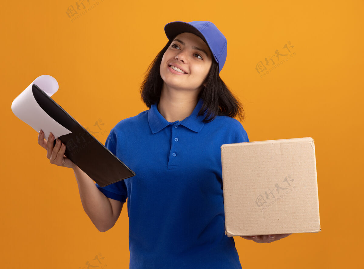 剪贴板身穿蓝色制服 戴着帽子的年轻送货员拿着空白页和纸板箱的剪贴板 站在橙色的墙上 面带微笑地看着我女孩举行微笑