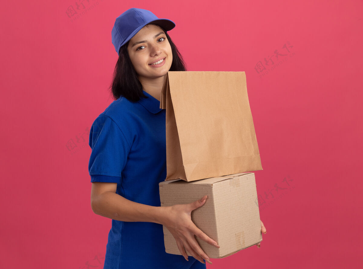 送货身穿蓝色制服 头戴鸭舌帽 手持纸包装和纸板箱的年轻送货女孩站在粉红色的墙上 脸上露出幸福的笑容包装女孩制服