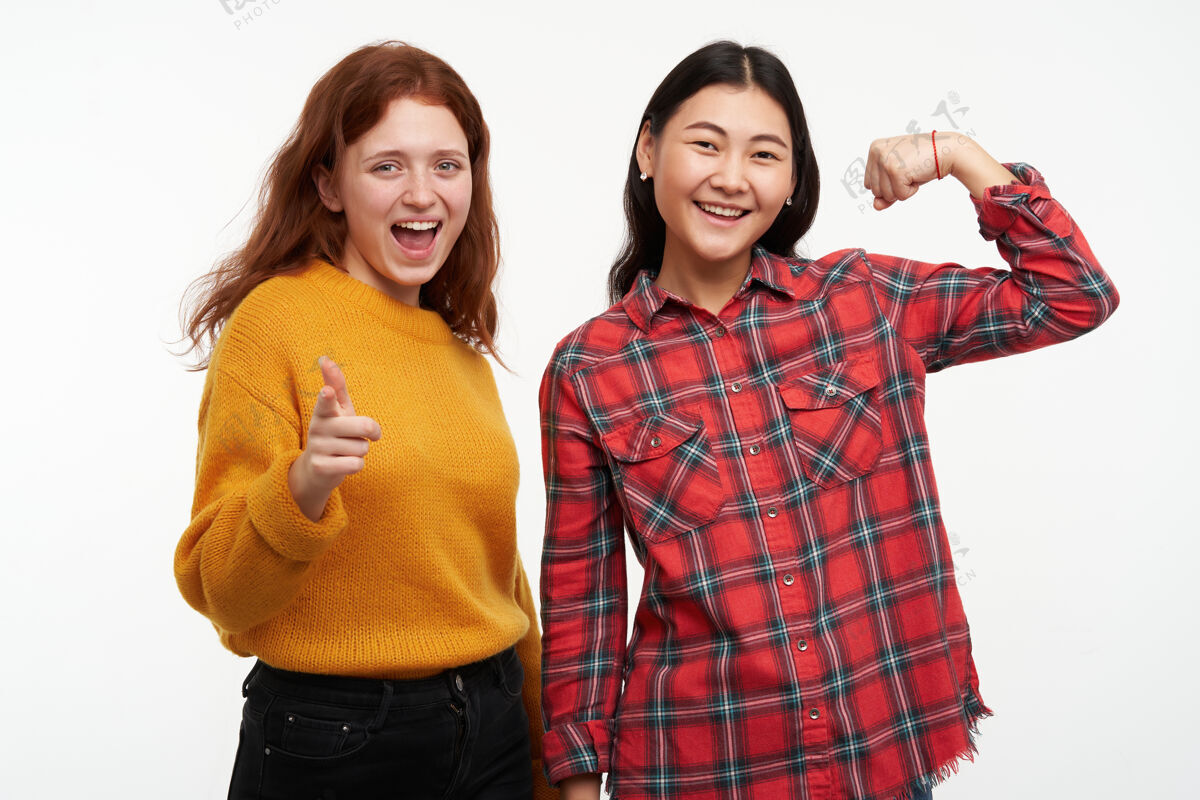 女性人和生活方式的概念两个快乐的女孩穿着黄色毛衣和格子衬衫指着你微笑和朋友显示肱二头肌隔离在白色的墙壁服装毛衣女人