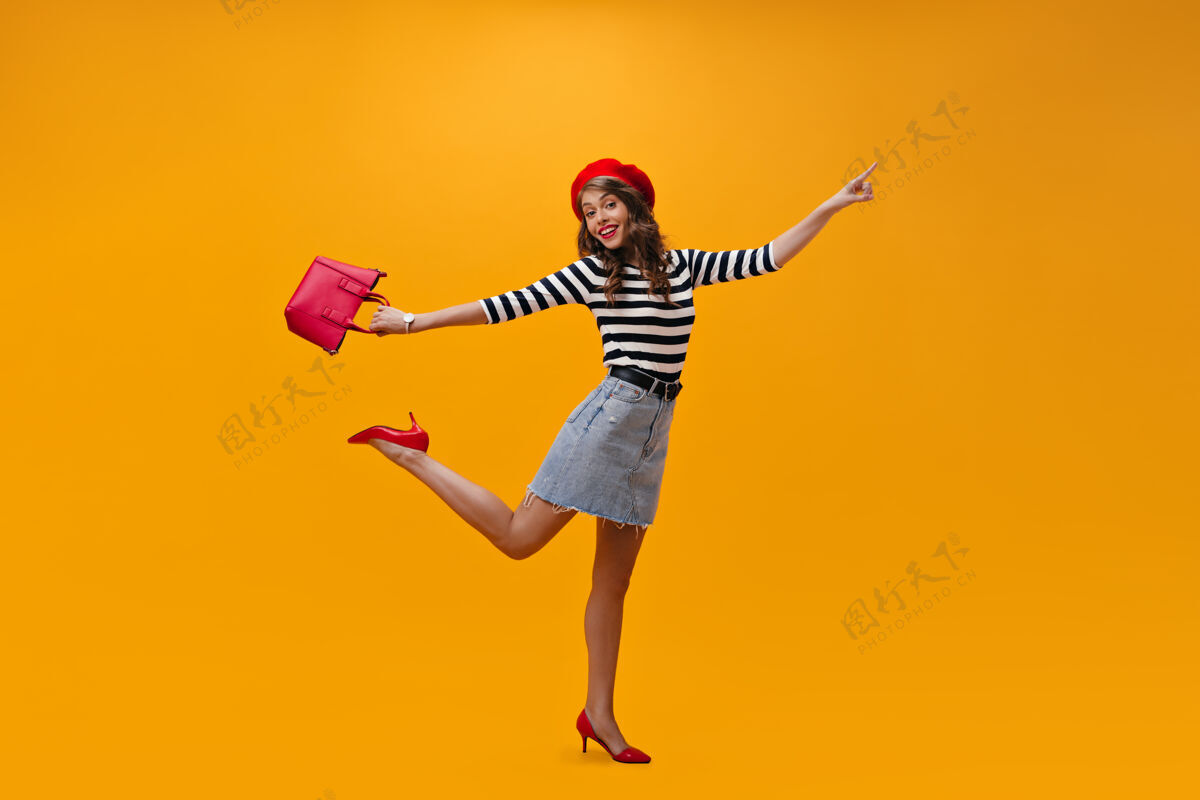 头发快乐的女士穿着条纹衬衫 红色贝雷帽跳跃橙色背景带手提包漂亮的年轻女士戴贝雷帽和时髦的鞋子摆姿势休闲脸女性