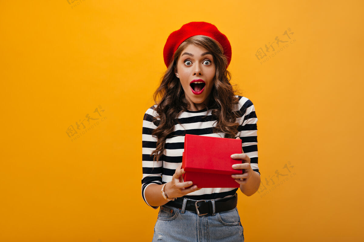 女孩卷曲的女人打开礼盒后看起来很震惊穿着红色贝雷帽和条纹衬衫的惊讶女孩看着镜头为礼物感到高兴脸女性姿势