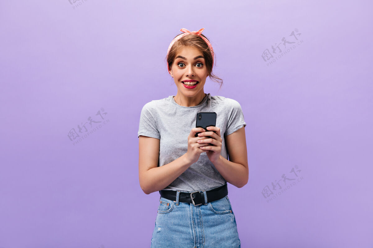 休闲女孩高兴地看着 手里拿着智能手机穿着灰色t恤和现代裙子的年轻女子惊讶地看着紫色背景下的相机时尚衬衫微笑