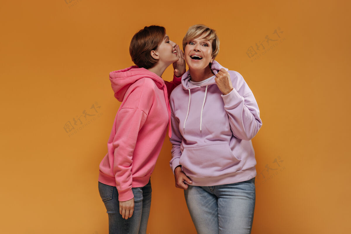 女士一个穿着淡紫色运动衫和牛仔裤的金发女人在橙色背景下听着孙女的秘密休闲老年人中等