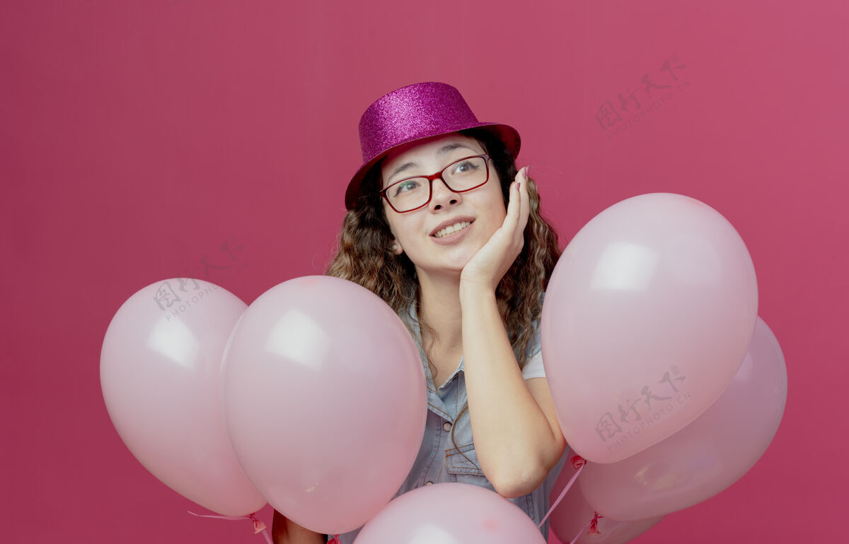 眼镜看着旁边高兴的戴着眼镜和粉色帽子的年轻女孩站在气球后面 把手放在粉色背景上孤立的脸颊上脸颊手戴