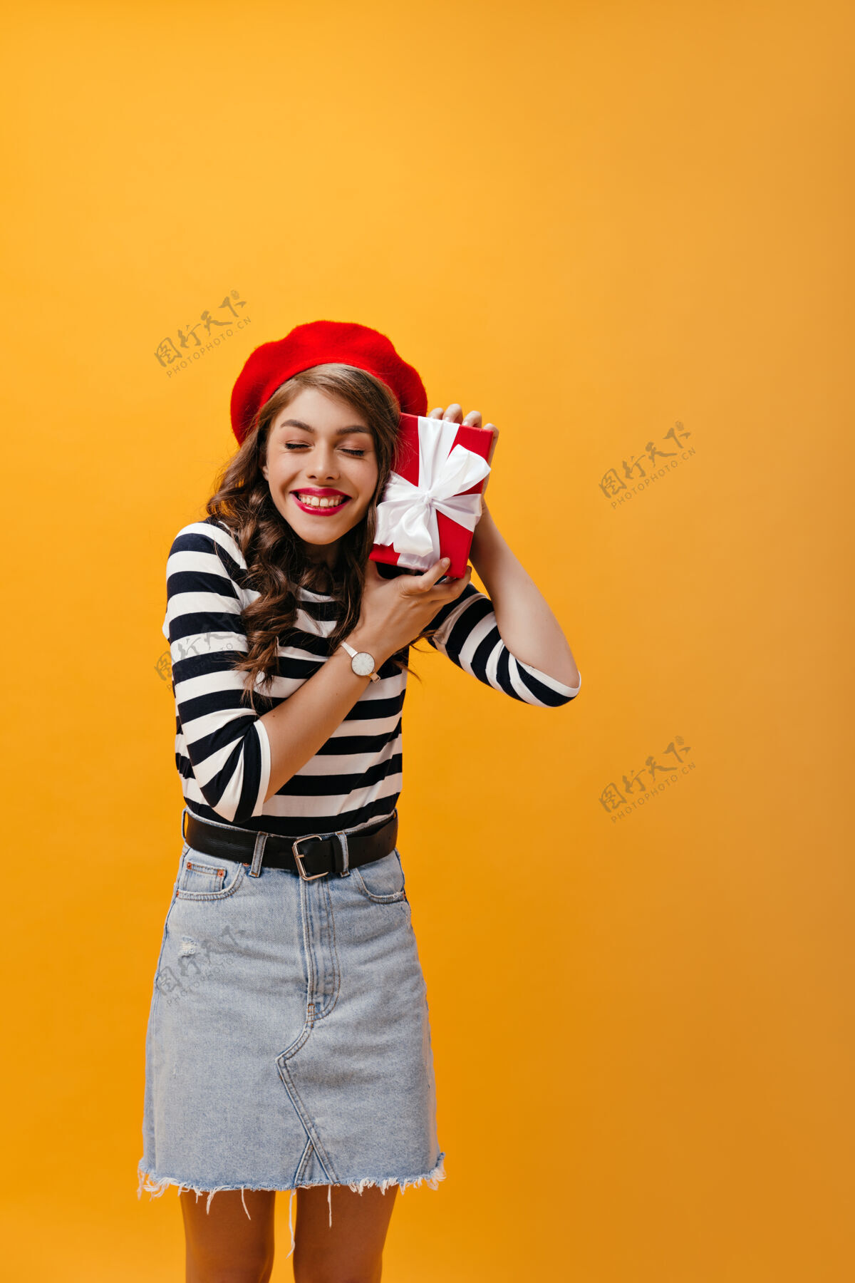 黄色穿着条纹衬衫和牛仔裙的快乐女孩拿着红色礼盒快乐的年轻女子 卷发 穿着现代服装 戴着手表微笑着成人空间表情