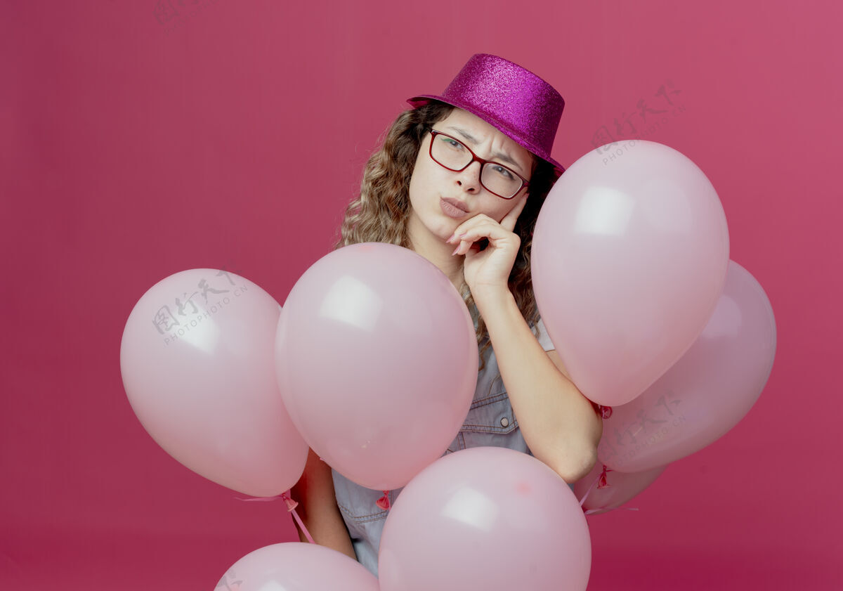 气球看着一旁想着戴着眼镜 戴着粉红色帽子的年轻女孩 手里拿着气球 把手放在脸颊上 隔离在粉红色的背景上穿思考手