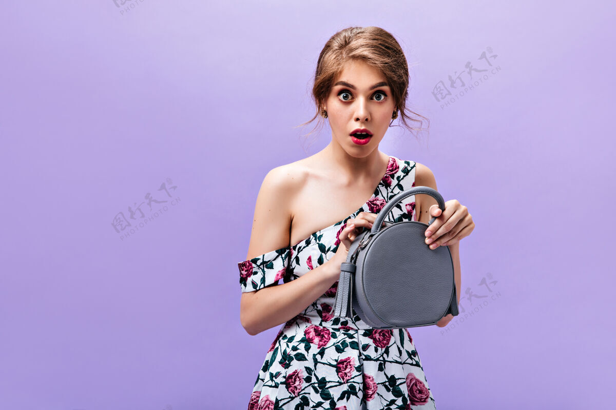 包一位惊讶的女士打开了她时尚的手提包一位身着花卉夏装的年轻漂亮女孩在与世隔绝的背景上拿着一个灰色的包女孩脸女人