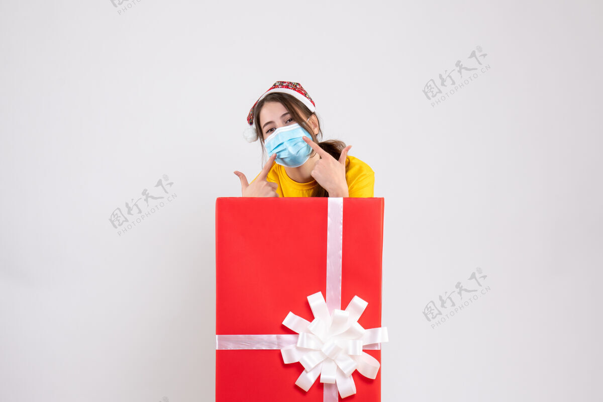 立场前视图可爱的圣诞女孩与圣诞帽指着她的面具站在大圣诞礼物后面丝带指向购物