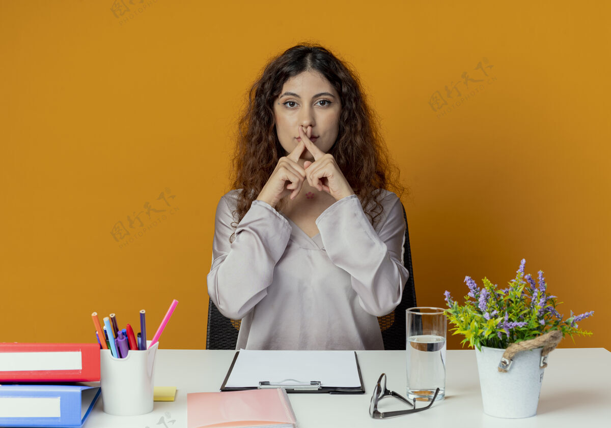 办公桌看着镜头 年轻漂亮的女上班族坐在办公桌前 手里拿着办公工具 在橙色的背景上显示出不孤立的姿态工人年轻人办公室