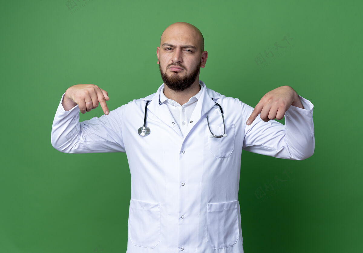 医疗身穿医用长袍和听诊器的自信的年轻男医生指着绿色背景上孤立的自己年轻自信听诊器