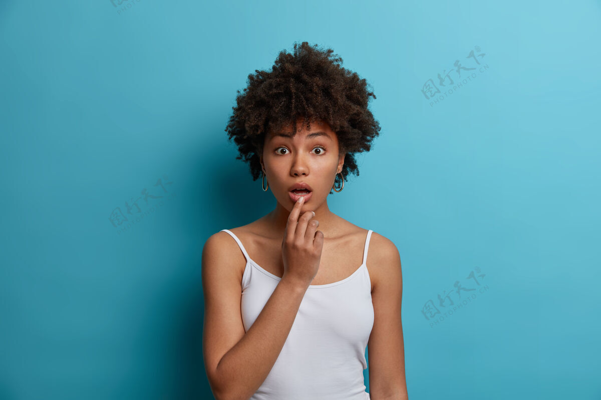 无语惊奇的非洲裔美国妇女的肖像从震惊中垂下下巴 听到令人惊奇的难以置信的消息 发现令人惊讶的缓和 听到谣言或坏消息 穿着白色背心 隔离在蓝色的墙上反应好奇卷发
