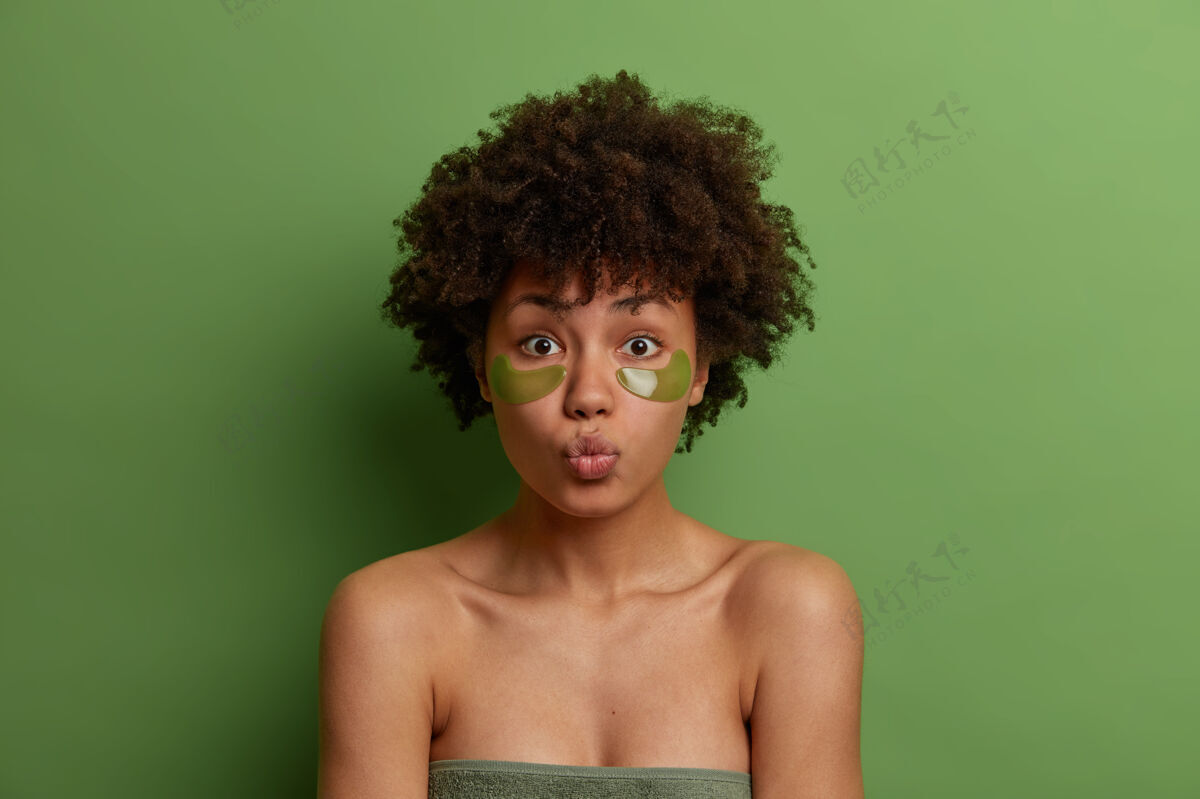 卫生面部护理概念可爱的年轻女性 非洲发型 眼睛下面有绿色的斑块 嘴唇圆润 毛巾裹着站着 自然美丽 在绿色的墙壁上摆姿势圈子毛巾卷曲