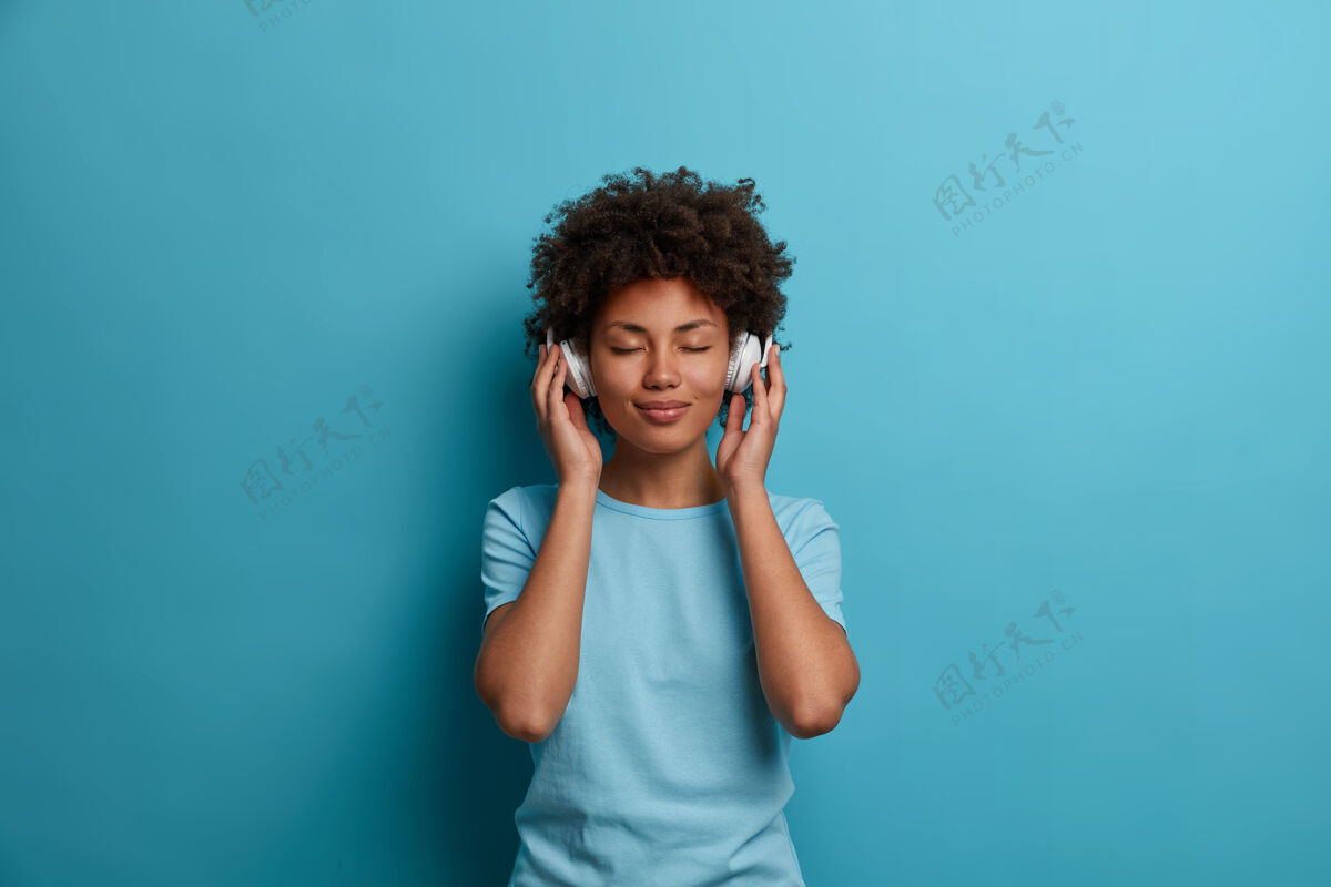 耳机满足的卷发美籍年轻女性有着无忧无虑的快乐心情 闭上眼睛 戴着耳机听音乐 穿着休闲的蓝色t恤 在室内摆姿势人 休闲 娱乐的理念满足青少年音频