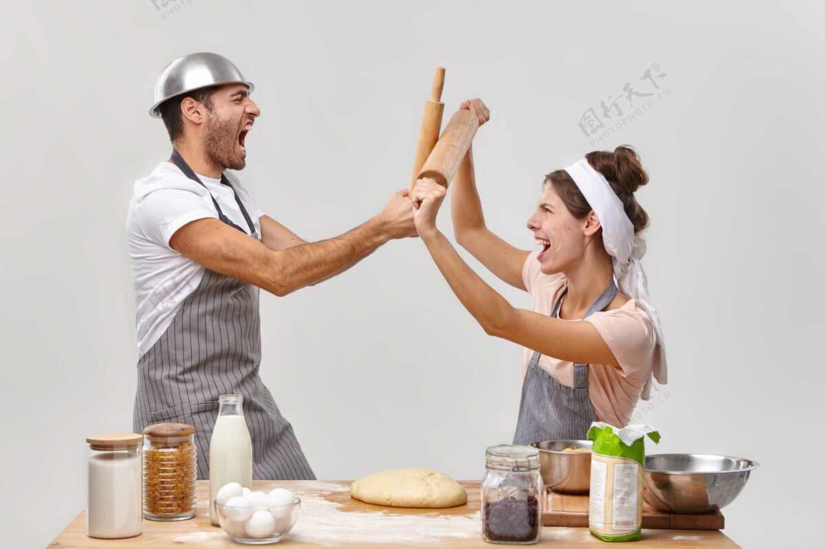 尖叫男女对手的横拍参与烹饪挑战 用木制擀面杖战斗 进行烹饪战斗 在面包房工作 做面团 在厨房对着白墙摆姿势愤怒面粉规则