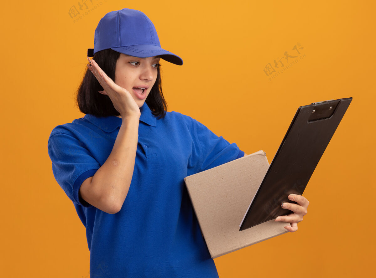 交货身穿蓝色制服 头戴鸭舌帽的年轻送货女孩站在橙色的墙上 手里拿着纸板箱和剪贴板 惊讶地看着它制服举行女孩