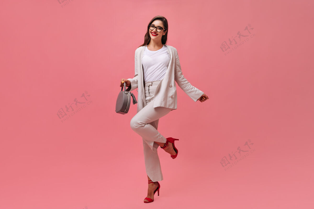 高跟鞋穿着米色套装的漂亮女人在粉色背景上愉快地摆姿势一个穿着白西装红鞋子 带着灰色手提包的开朗女孩在微笑时尚站立女孩