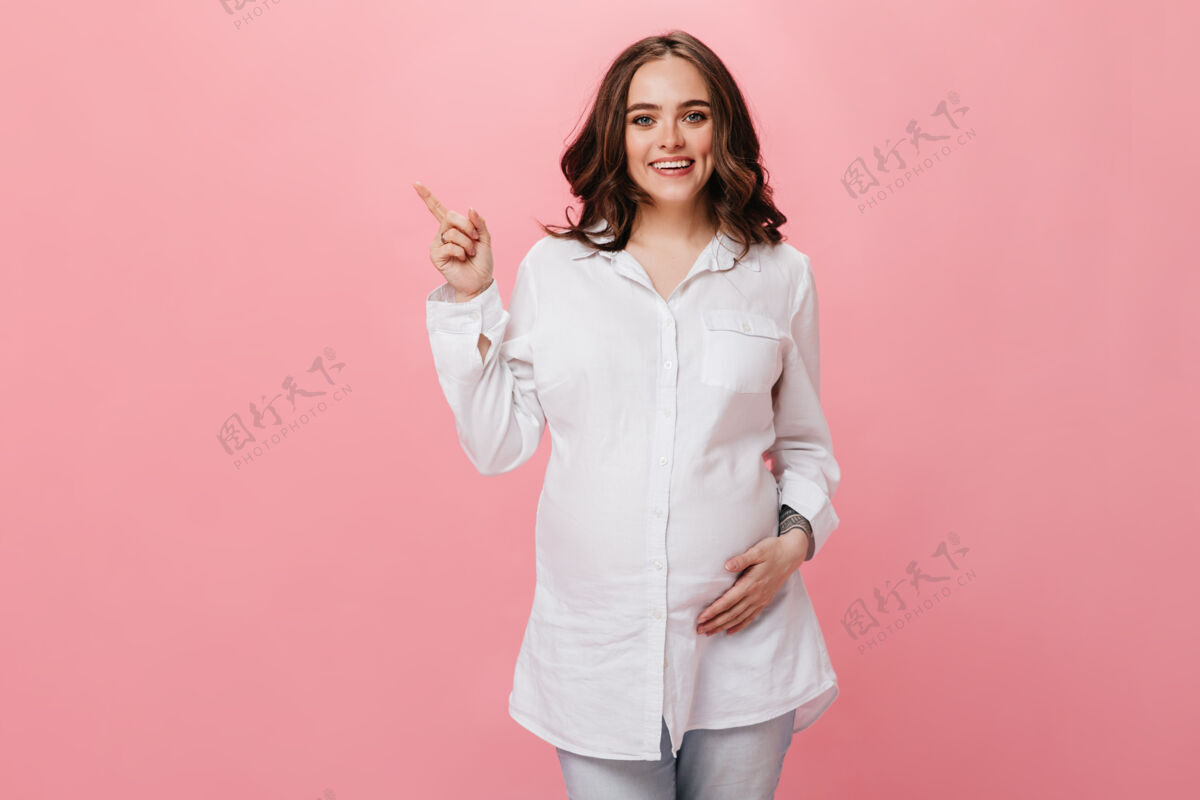 肚子幽默的孕妇抚摸着肚子 微笑着身穿白衬衫的黑发女孩指着孤立的粉红色背景上的文字微笑白衬衫腹部