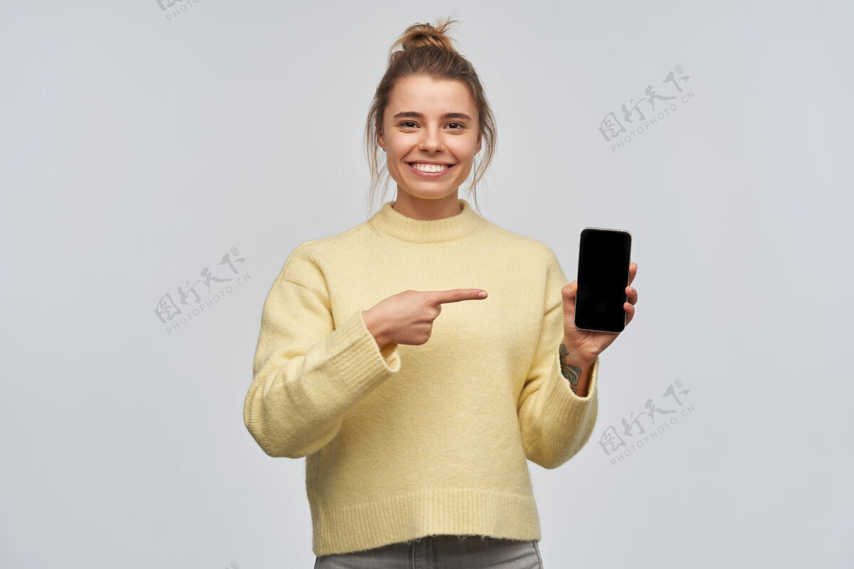 手机照片中的美女金发束成发髻 身穿黄色毛衣 指着手机屏幕 复制空间面带微笑看着镜头 隔着白色的墙壁休闲模特情绪
