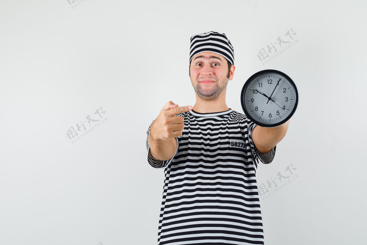 肖像穿着条纹t恤的年轻男子 戴着帽子指着时钟 看上去很高兴 正对着前方优雅英俊脸