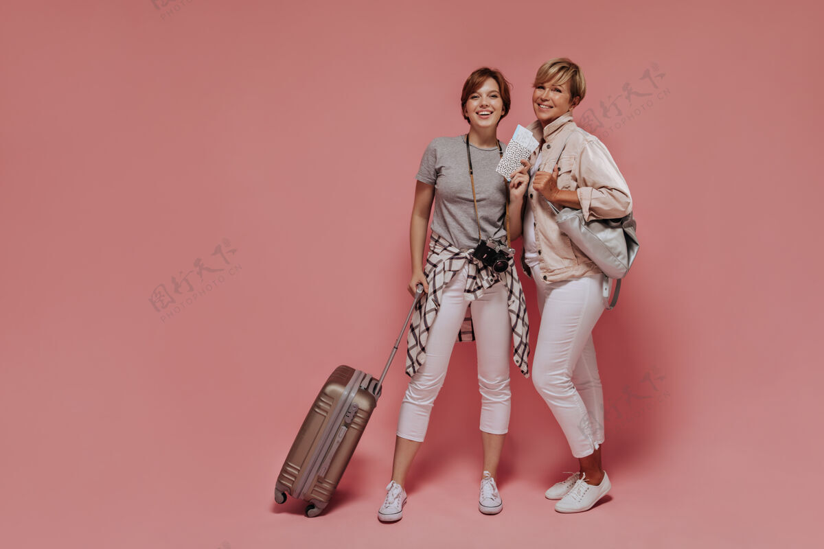 灰色一个快乐的女孩 深色头发 穿着浅色裤子和灰色t恤 手里拿着手提箱 票和相机 在粉色背景上和微笑的女人合影手提箱年龄粉色