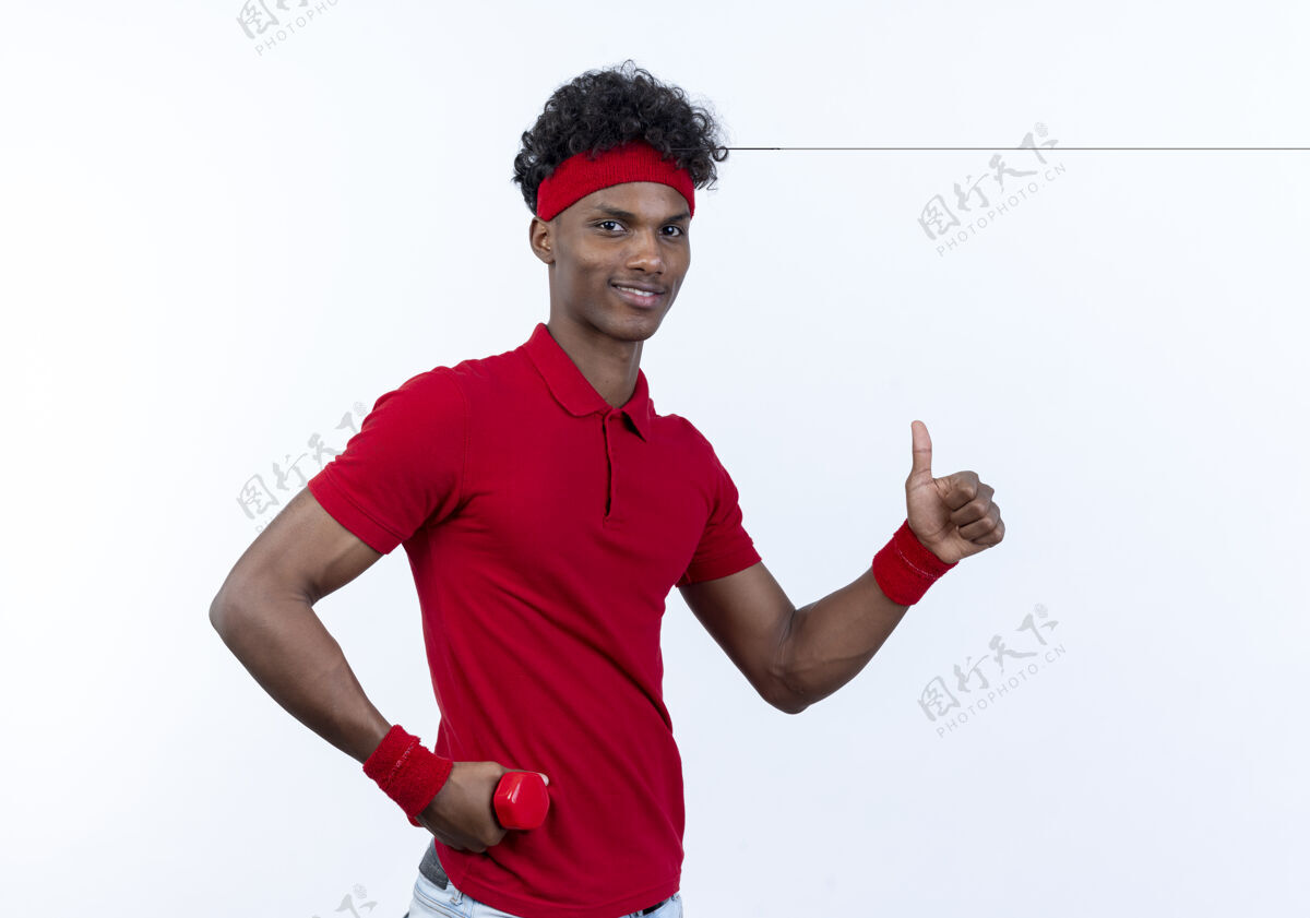 年轻高兴的年轻黑人美国运动男子戴着头带和手环举着哑铃臀部他的大拇指上孤立的白色背景手握头带拇指