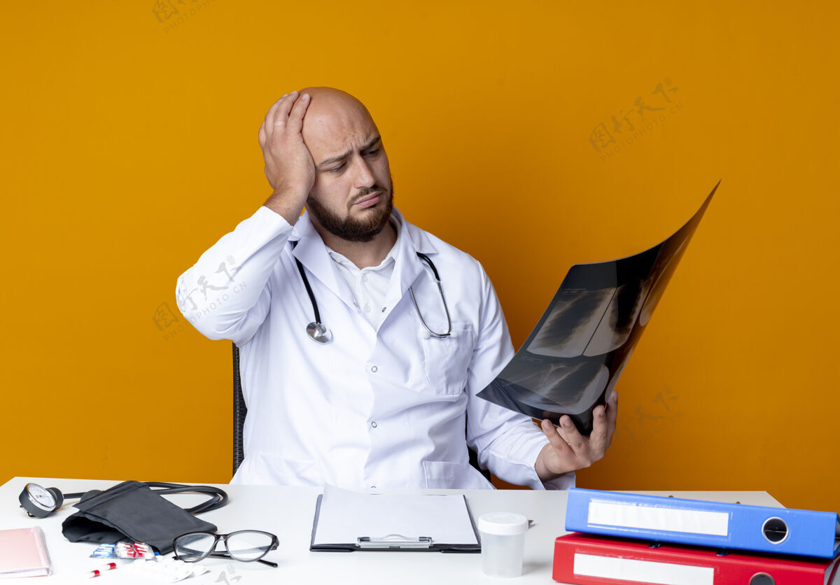 持有困惑的年轻秃头男医生穿着医用长袍和听诊器坐在办公桌旁 手持医疗工具 看着隔离在橙色背景上的x光片长袍坐着秃头