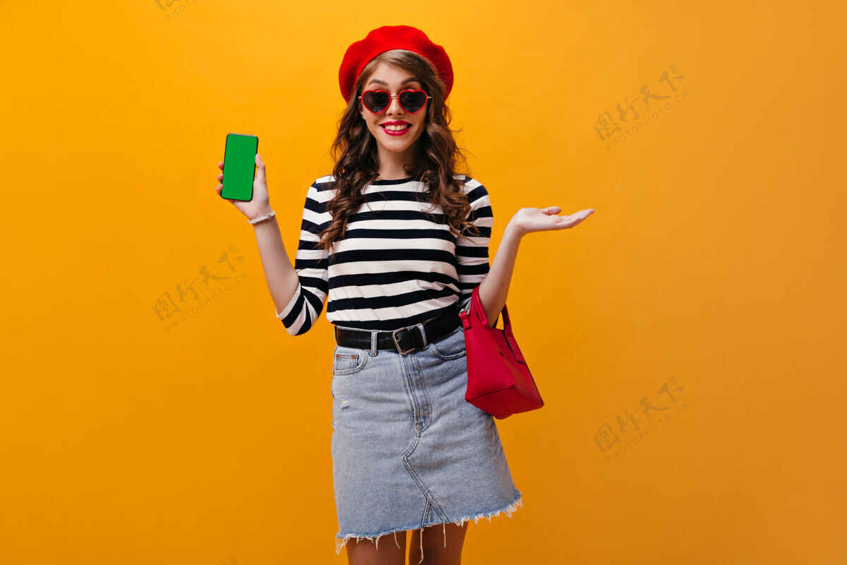表情戴着墨镜和红色贝雷帽的漂亮女人展示智能手机穿着粗斜纹棉布裙子 黑色宽腰带的迷人女孩在孤立的背景下微笑脸复制紫色