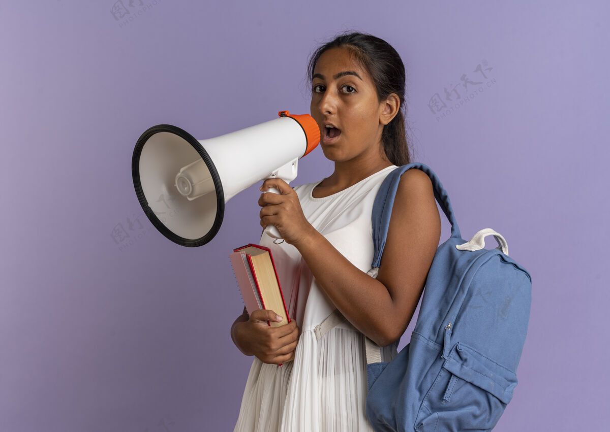 说话看着镜头 一个背着书包 拿着书的年轻女学生在孤立的紫色背景下用扩音器讲话年轻人包女学生