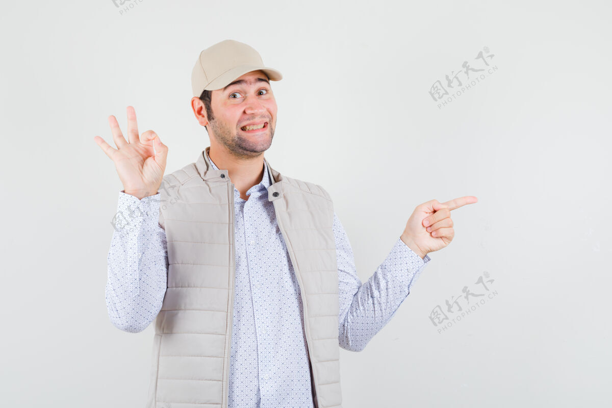 男性穿着米色夹克和帽子的年轻人显示ok标志 食指指向右边 看起来很高兴 正面视图脸指人