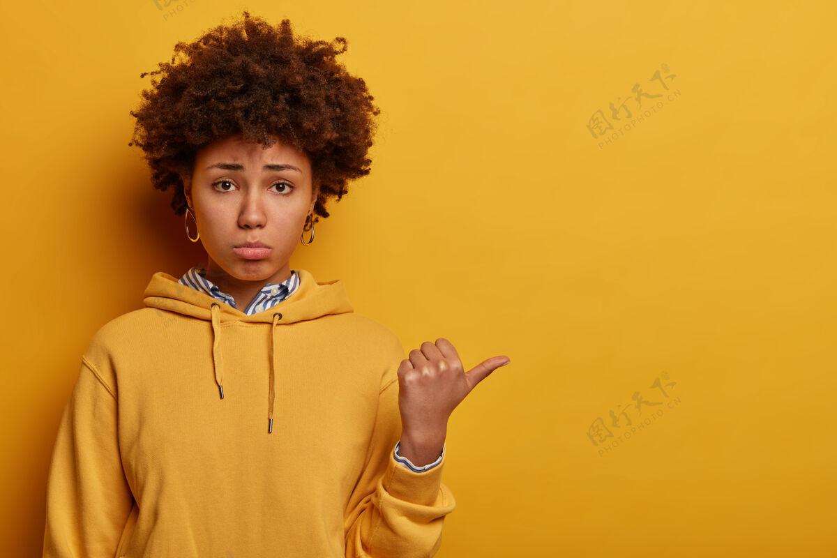 负面不满意的黑皮肤女人的肖像用拇指指向别处 给出负面反馈 对令人失望的事情做出反应 抱怨和抱怨 穿着休闲连帽衫 隔离在黄色的墙上反应情绪建议