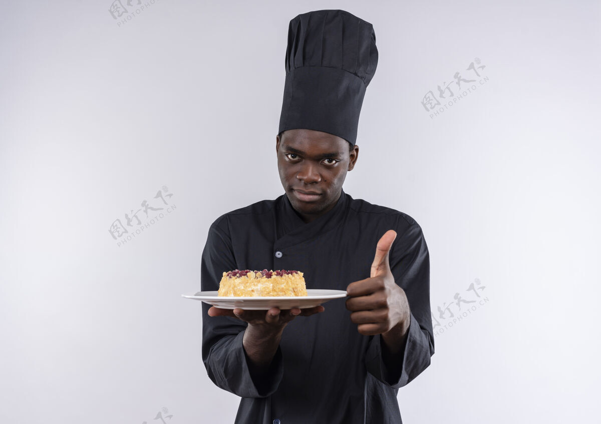 制服年轻自信的非洲裔美国厨师身着厨师制服 把蛋糕放在盘子上 竖起大拇指放在白色的空白处持有空间厨师