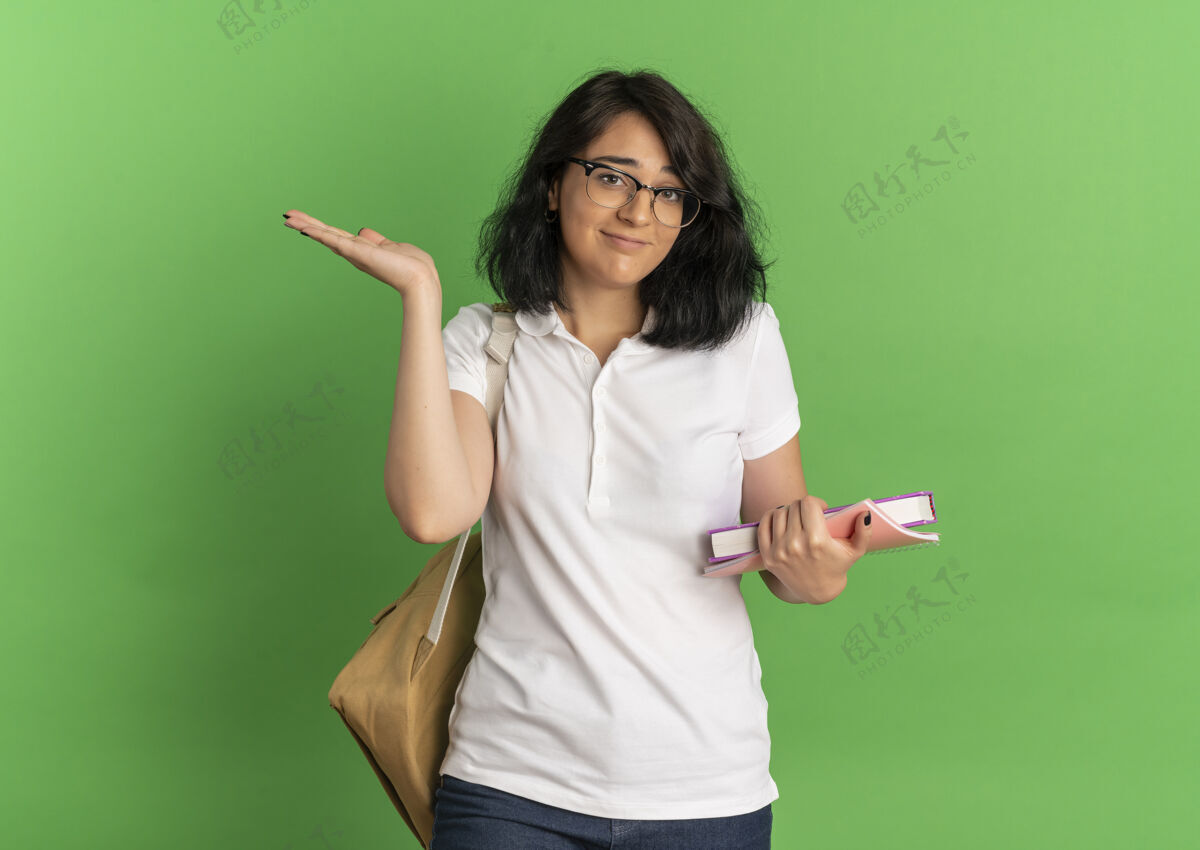 年轻年轻迷茫漂亮的高加索女学生戴着眼镜 背着书包拿着书 在绿色和空白处举手绿色女学生背