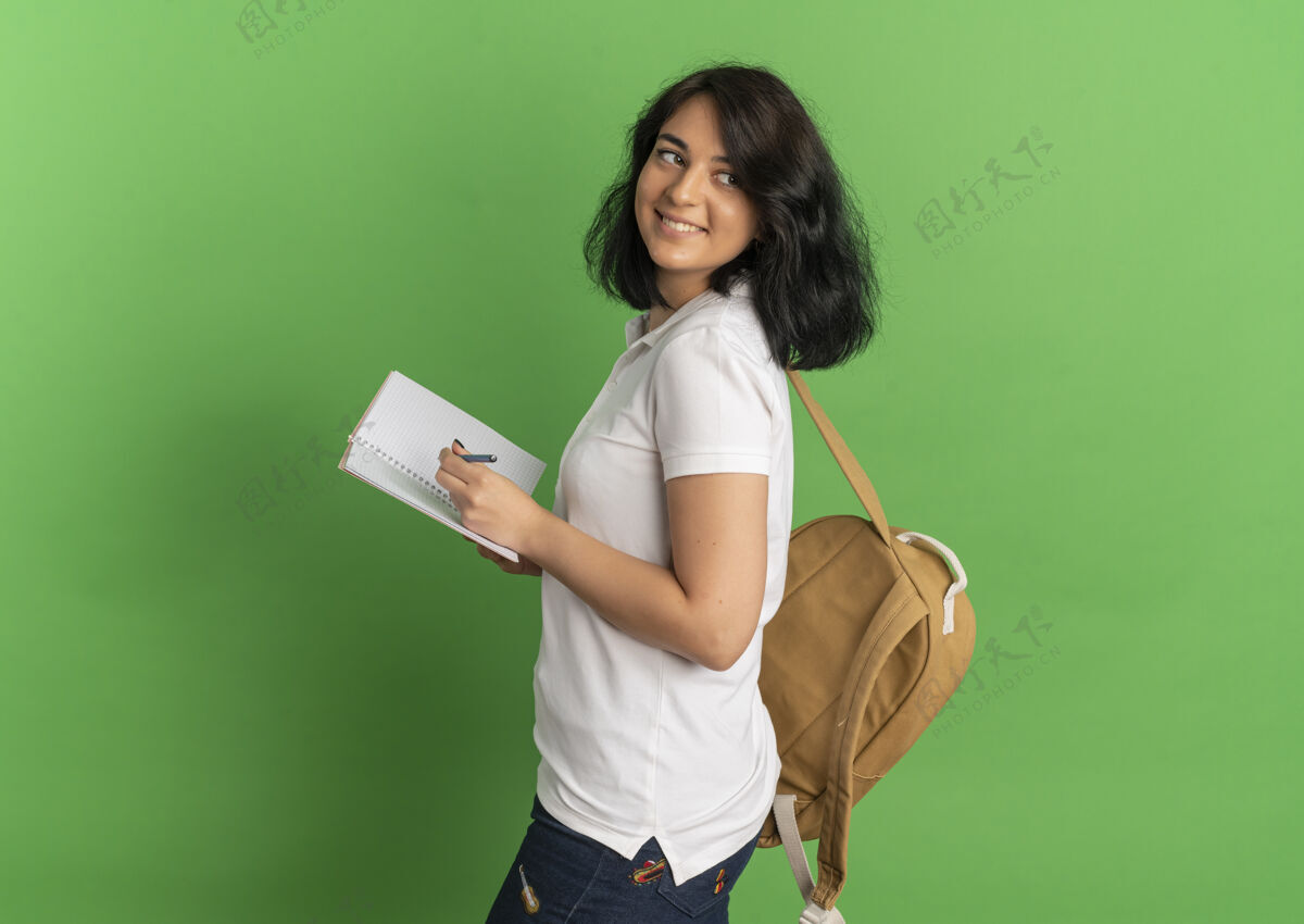 女学生年轻的高加索女孩 面带微笑 戴着眼镜 背着书包 站在一边 拿着笔记本 放在绿色的空间里年轻空间眼镜
