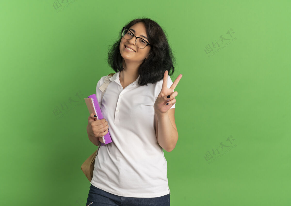 年轻年轻笑容可掬的高加索女学生戴着眼镜 背着书包 手举胜利的手势 手里拿着书 绿色上有复印空间标志穿眼镜